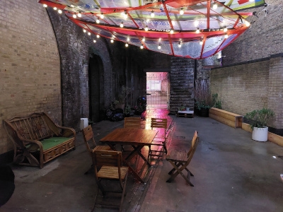 Lounge studio green panel wall with adjoining beauty salon and kitchen. Spiral staircase and door to garden.
Garden studio with table, chairs, benches and festoon light. The lounge studio can be converted to green screen.